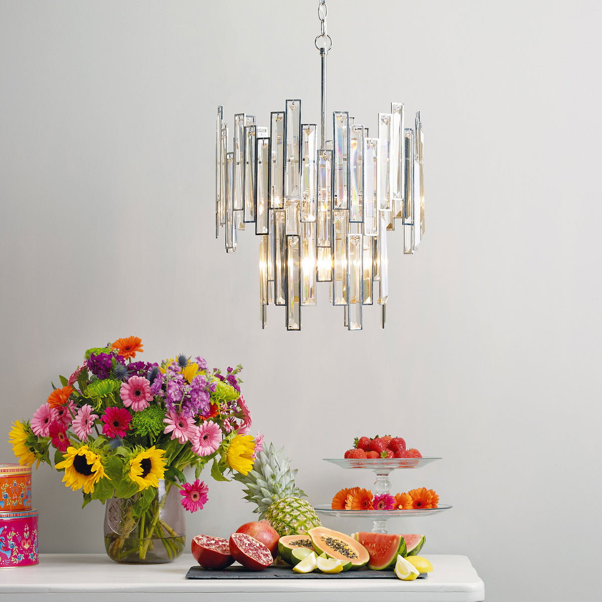A modern crystal chandelier hangs above a table with a colorful flower bouquet and fresh fruit.