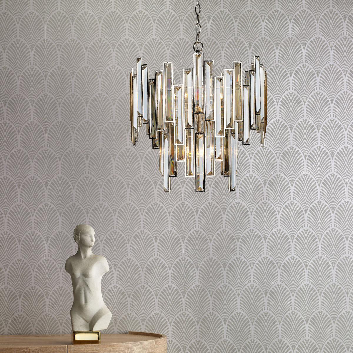 A crystal chandelier hangs from the ceiling, with a white bust on a wooden table below it. The wall has a gray art deco pattern.