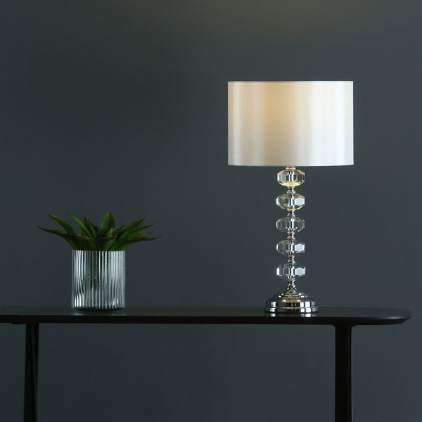A modern table lamp with a crystal base and white shade next to a potted succulent on a dark table.