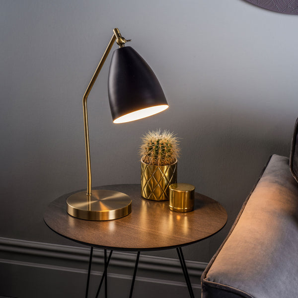 A black and gold table lamp illuminates a cactus in a gold pot and a gold candle on a round wooden table.