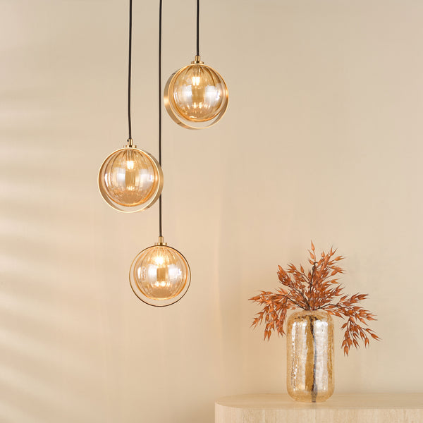 Three golden glass pendant lights hang from the ceiling, illuminating a textured beige wall. A vase with dried foliage sits on a light-colored table.