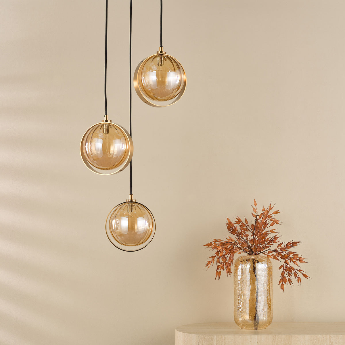 Three amber glass globe pendant lights hang above a textured gold vase with dried foliage.