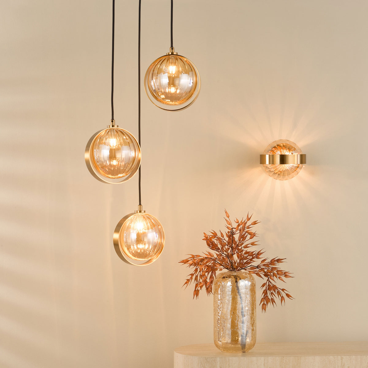 Three amber glass pendant lights and a matching wall sconce illuminate a beige wall. A vase with dried plants sits below.
