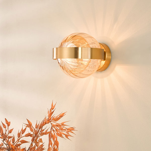A modern wall sconce with a spherical amber glass shade and a brass band, casting warm light upwards.