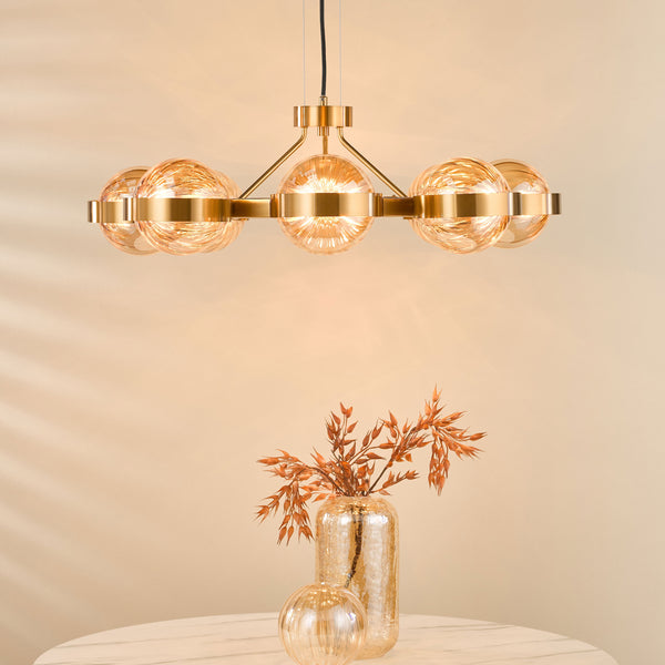 A gold chandelier with five textured glass globes illuminates a marble table with a vase of dried plants.