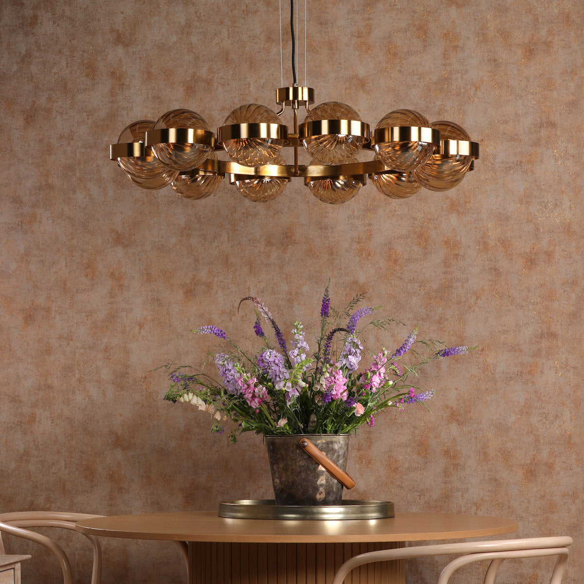 A gold chandelier with glass globes hangs above a wooden table with a bucket of purple and pink flowers.