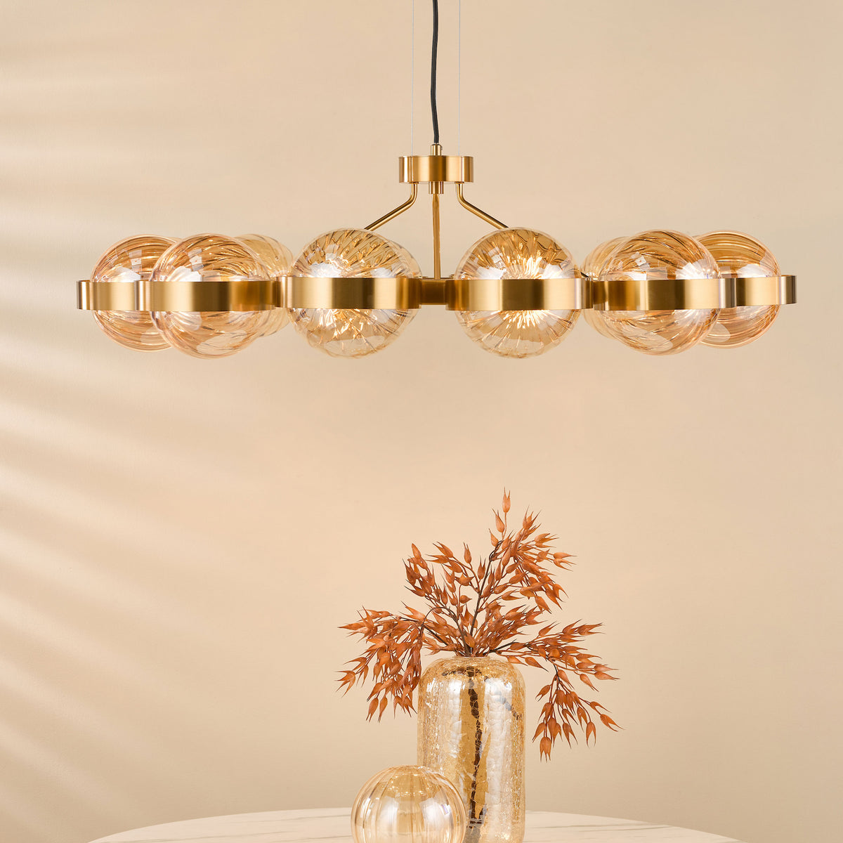 A golden chandelier with eight amber glass globes hangs above a table with a tall, cracked gold vase holding dried orange foliage and a smaller, ribbed amber glass sphere.