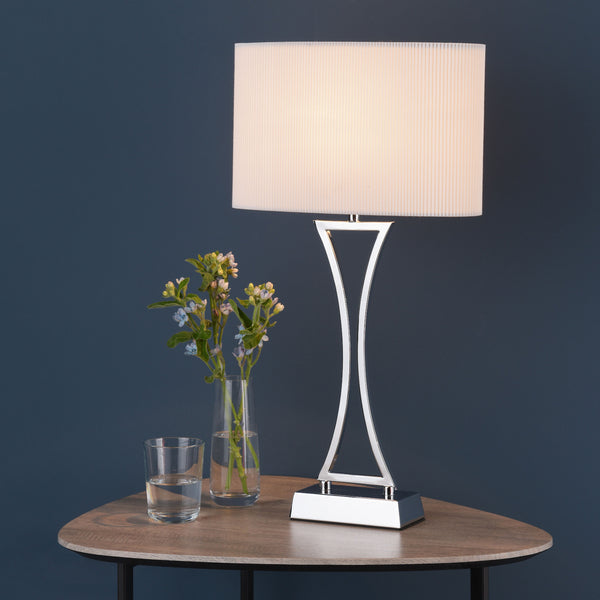 Table lamp with a white pleated shade and a chrome base on a wooden table with a vase of flowers.