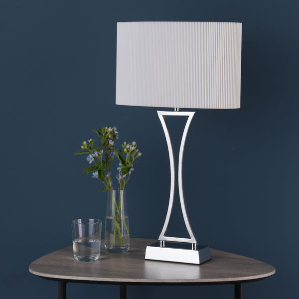 Modern table lamp with a white pleated shade and a chrome base, next to a vase of blue flowers and a glass of water.