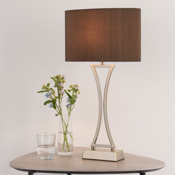 A modern table lamp with a brown shade and a unique base, next to a vase of flowers and a glass of water.