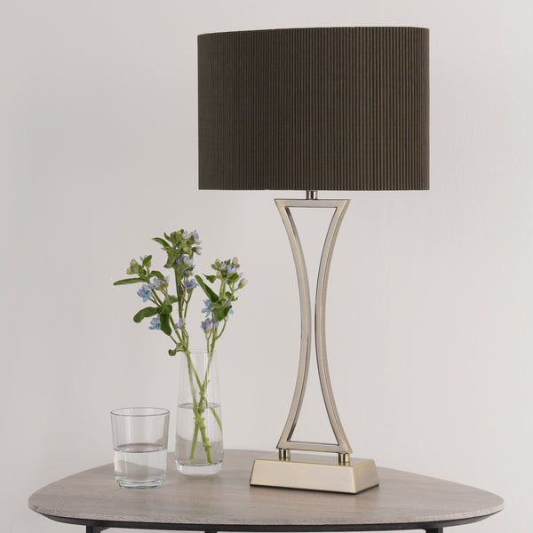 A modern table lamp with a dark pleated shade and a metallic base, next to a vase of blue flowers and a glass of water.