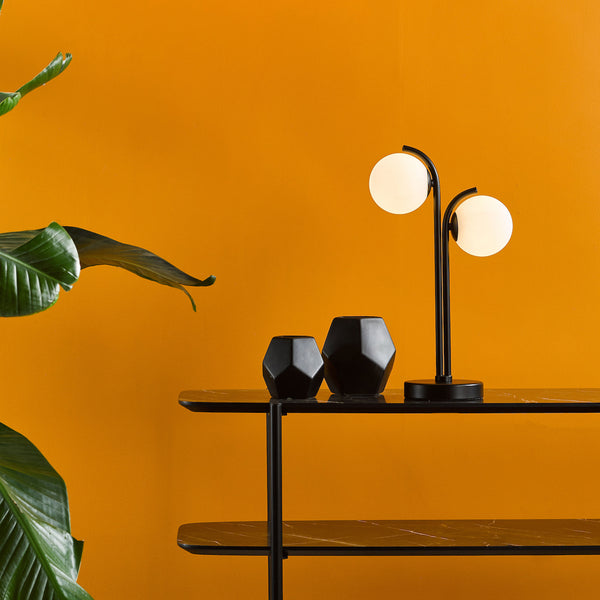 A black table lamp with two spherical lights, two black geometric vases, and a green plant against an orange wall.