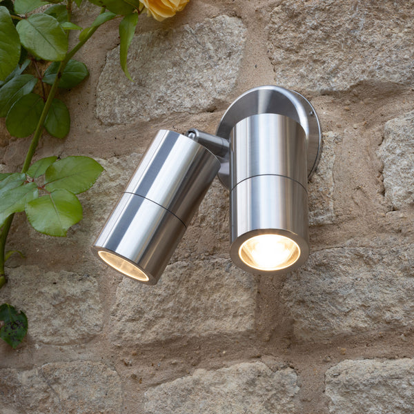 Two brushed stainless steel outdoor spotlights illuminate a stone wall with warm light, a rose vine visible.