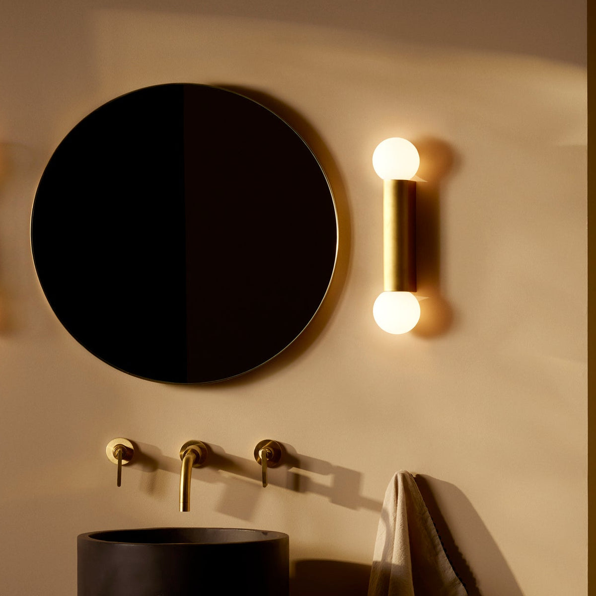 Round mirror, brass wall light, and black basin with brass taps