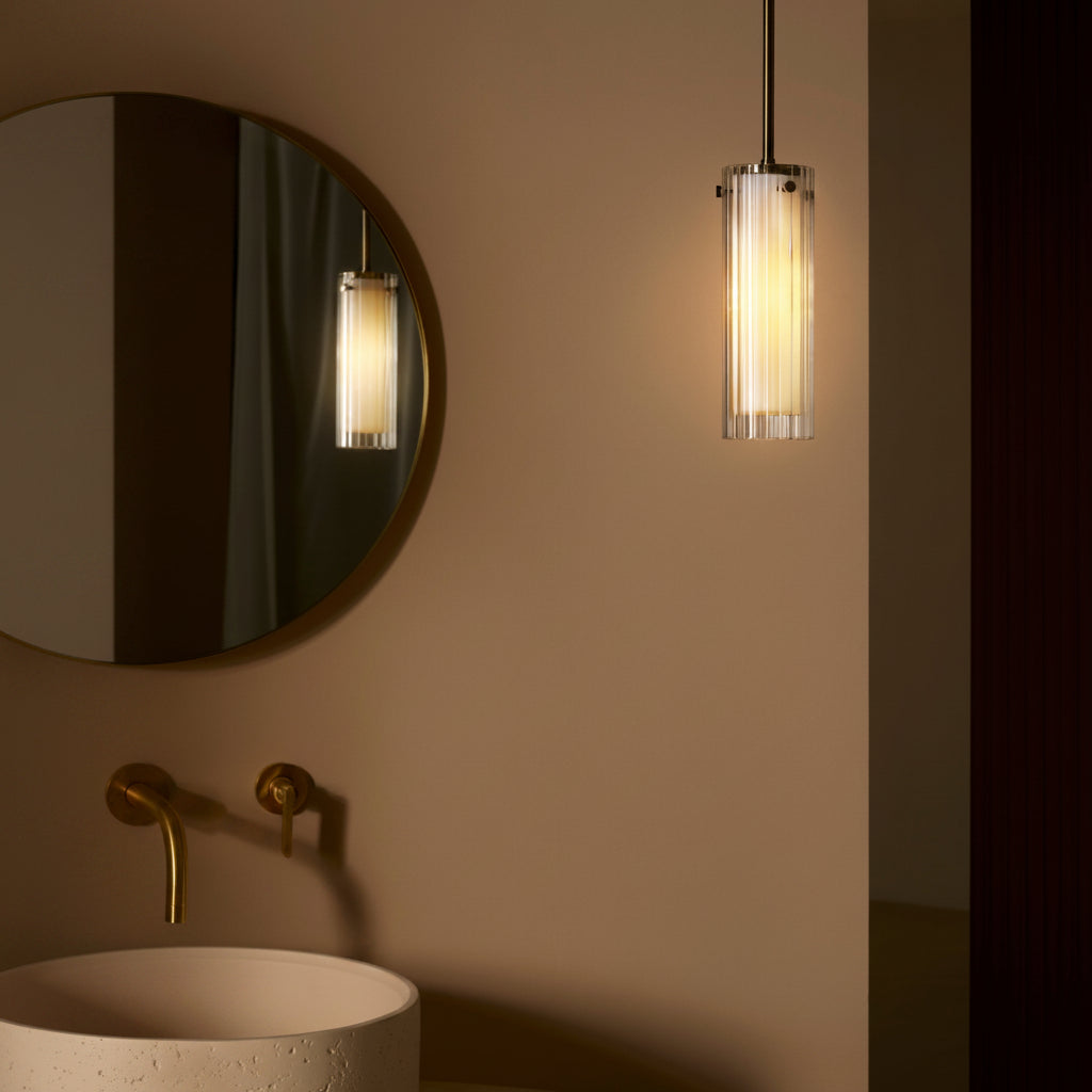 A modern bathroom with a round mirror, a concrete sink, brass taps, and a fluted glass pendant light