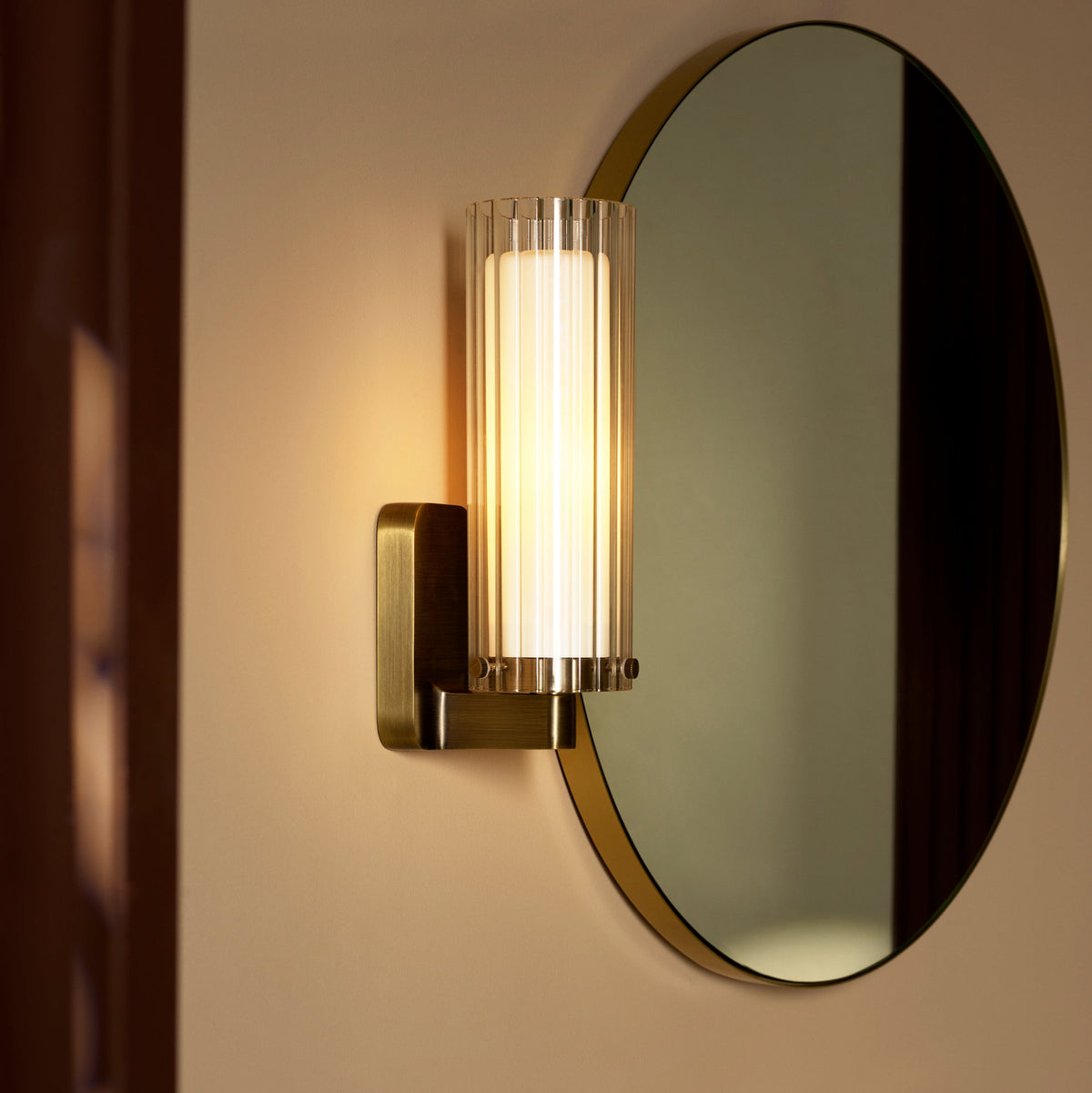 Wall-mounted cylindrical light fixture with a ribbed glass shade and a brass base next to a round mirror