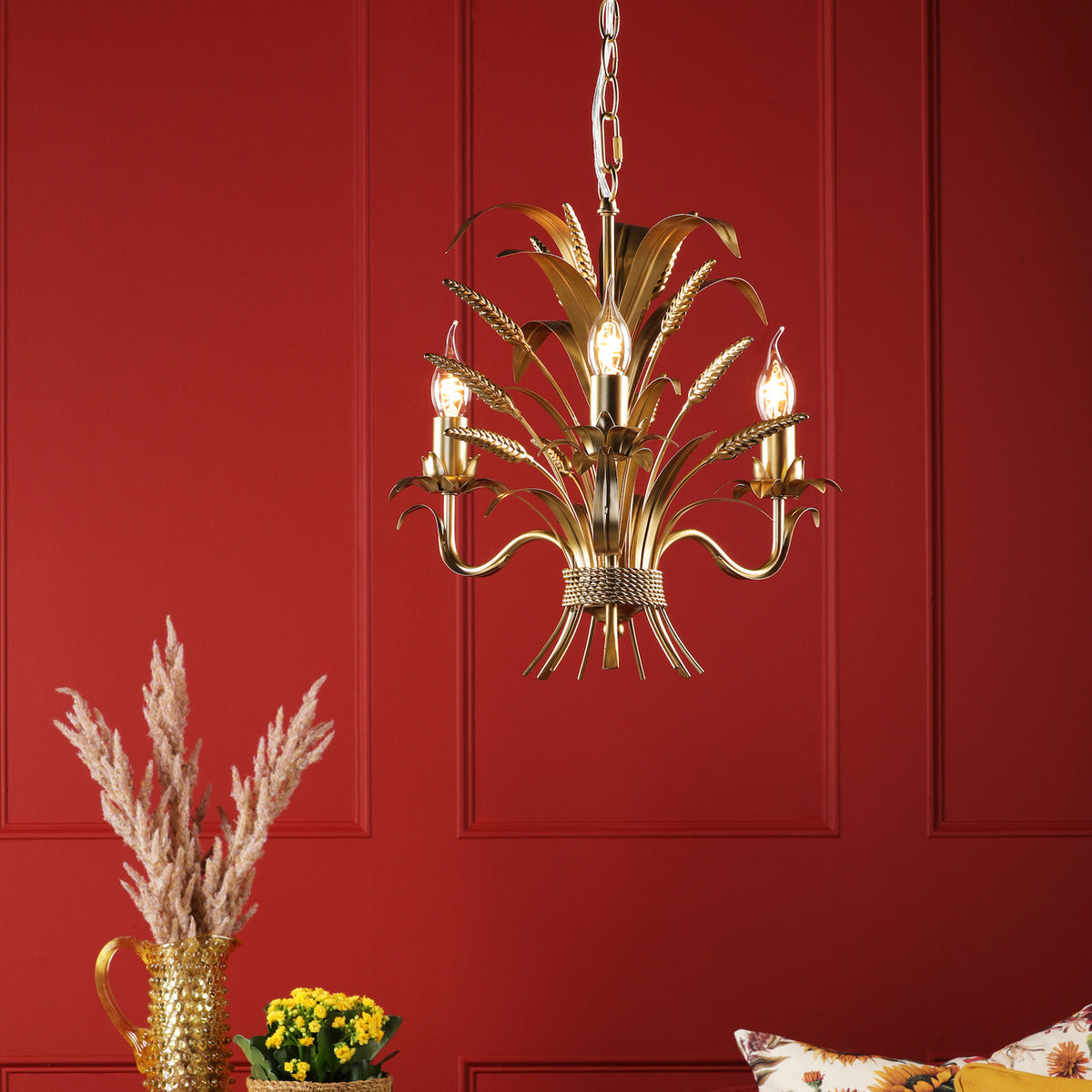 Gold chandelier with wheat stalk design and three candle-style bulbs against a red wall