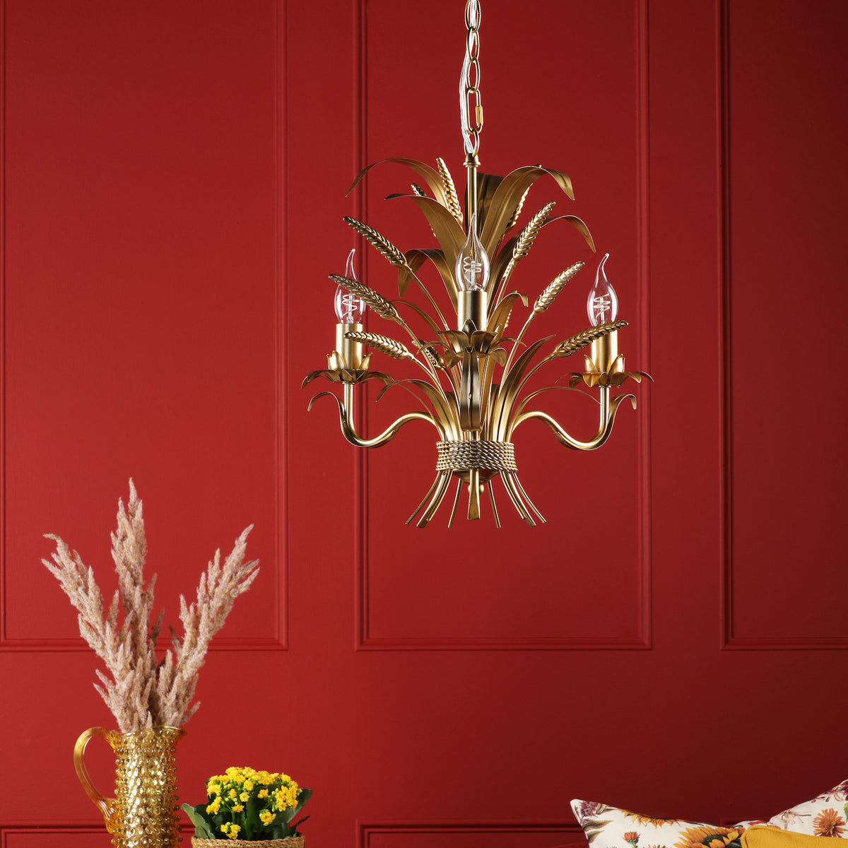 Gold chandelier with wheat stalk design and three exposed bulbs against a red paneled wall