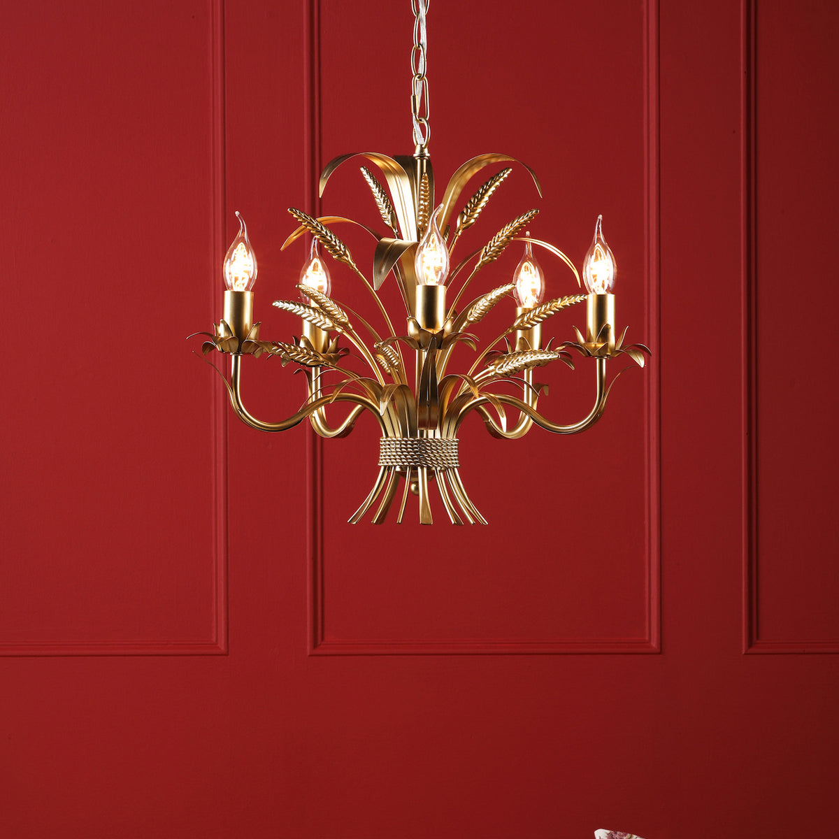 Gold chandelier with five candle-shaped bulbs and wheat sheaf details against a red paneled wall