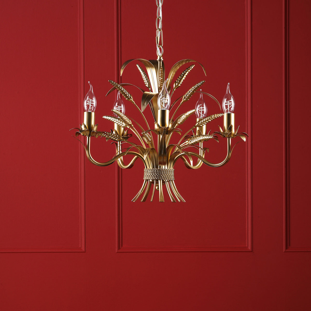 Gold chandelier with five candle-style bulbs, featuring decorative wheat sheaf designs, against a red wall
