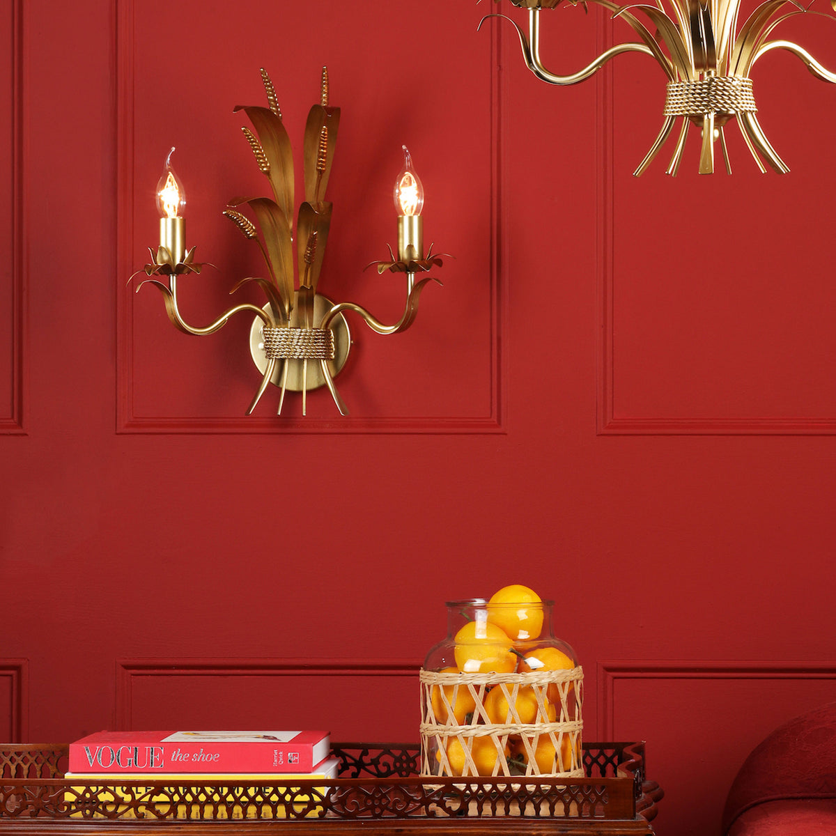 A gold wall sconce with two lights and a gold chandelier against a red wall