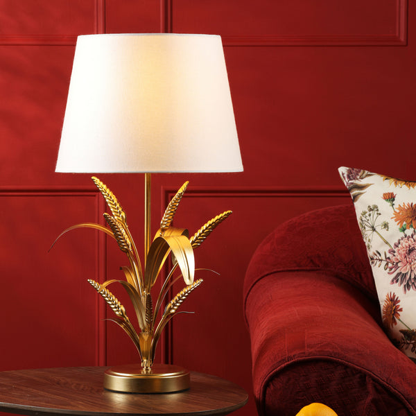 Gold wheat stalk table lamp with a white shade on a wooden table next to a red sofa and floral cushion