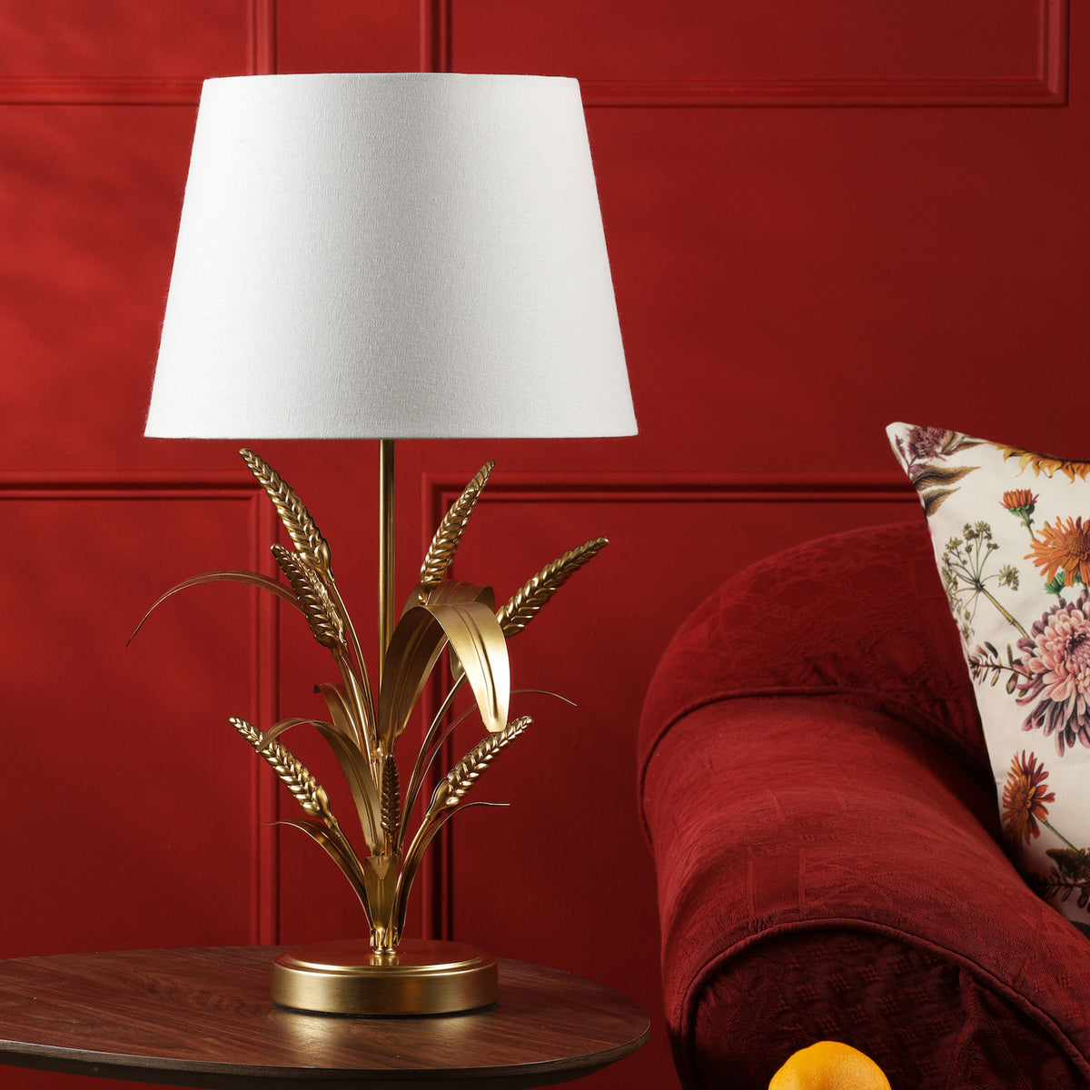 Gold wheat stalk table lamp with a white shade on a wooden table next to a red sofa