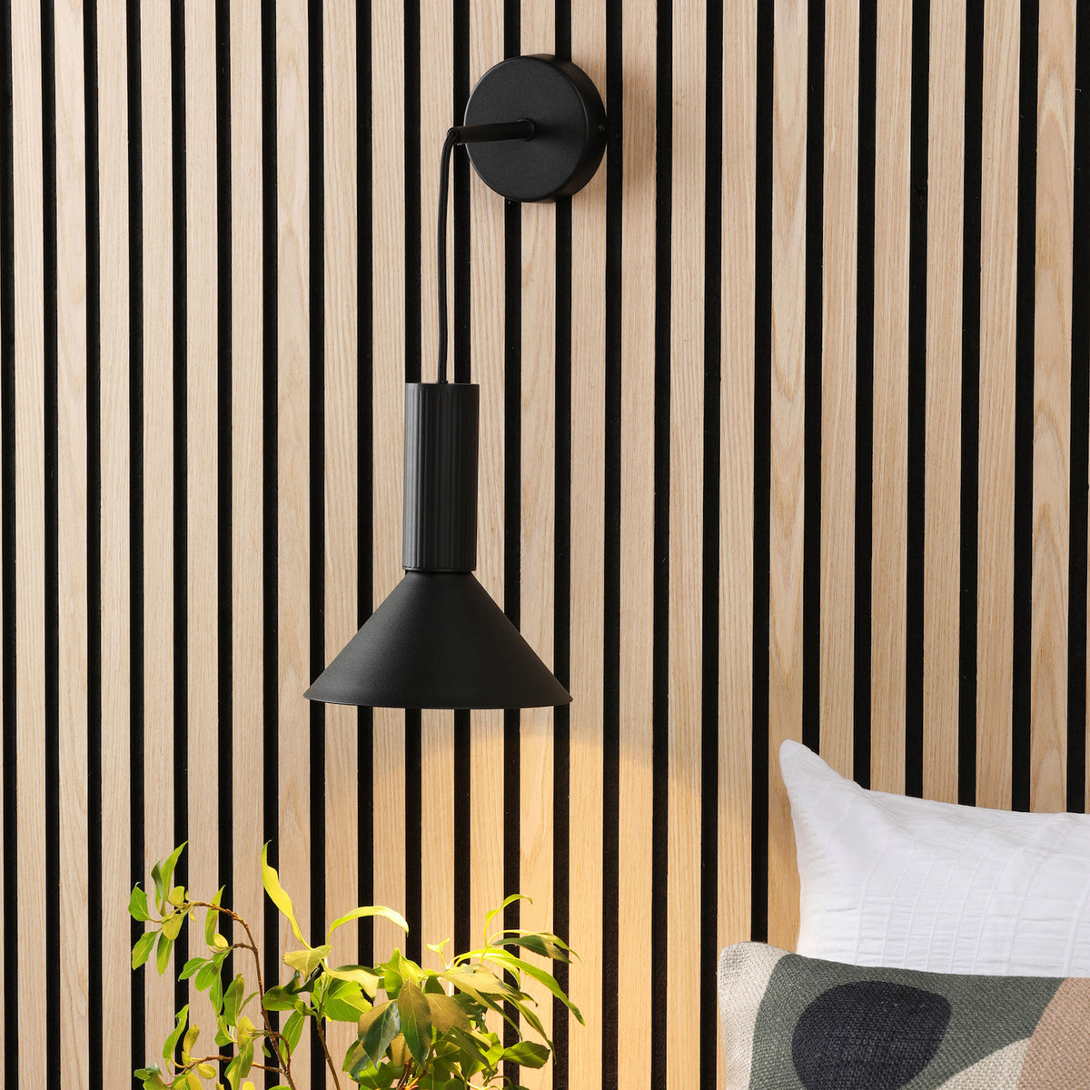 Black wall lamp on a wooden slatted wall, with a plant and a pillow in the foreground