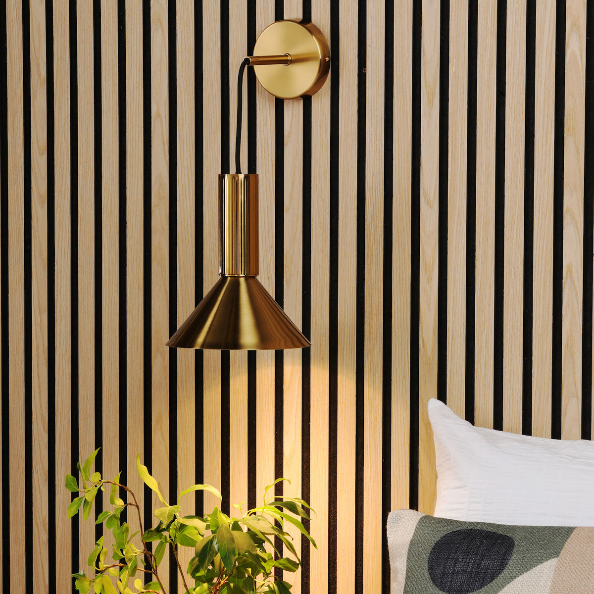 Brass wall lamp with a conical shade, illuminating a plant and a bed against a slatted wood panel wall