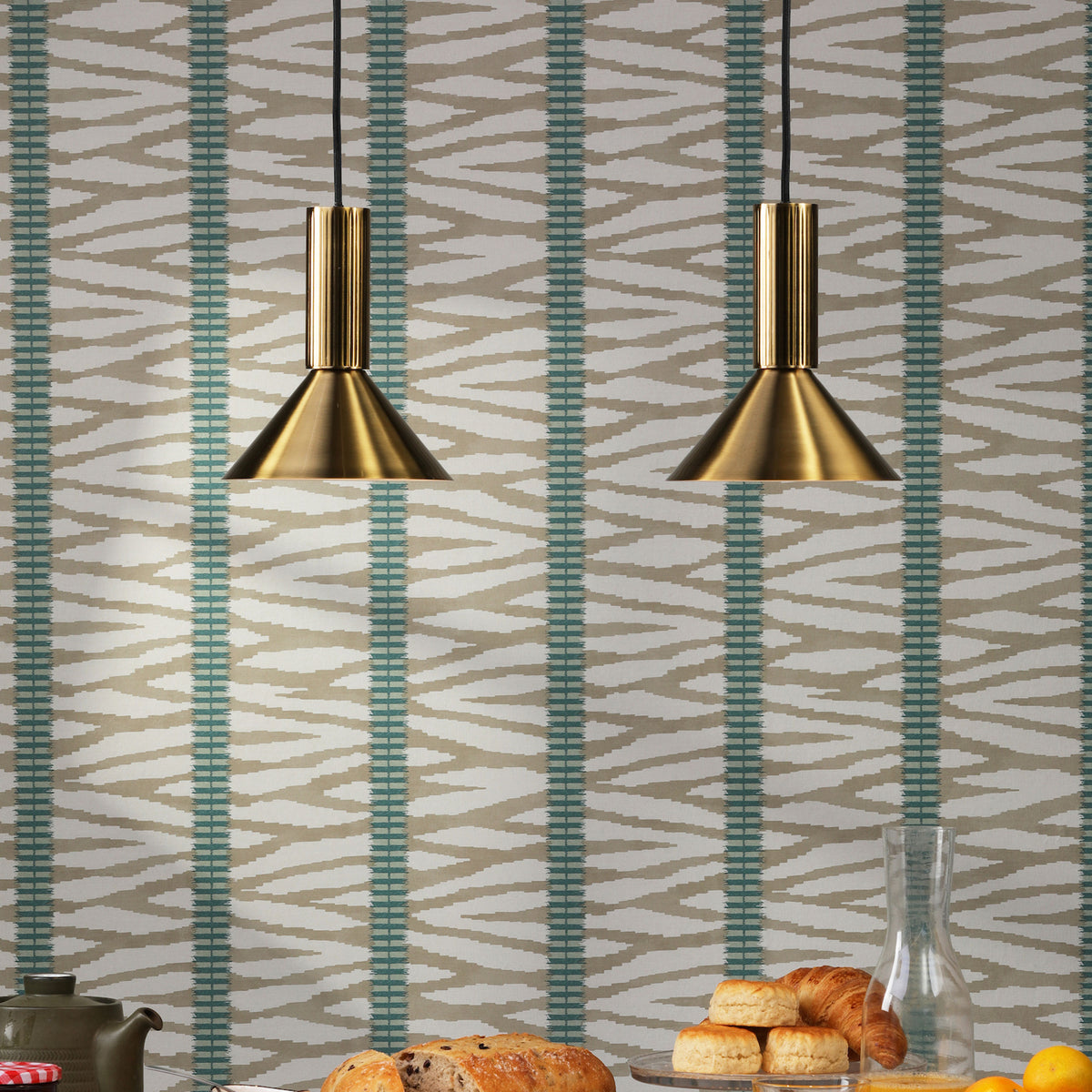 Two brass pendant lights hang in front of a patterned wallpaper with breakfast items on a table below