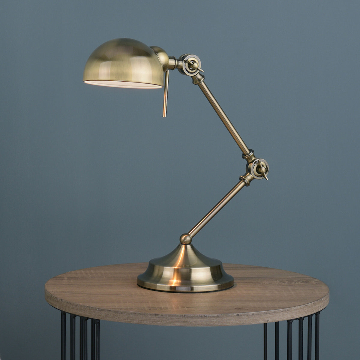 Adjustable brass desk lamp with a domed shade, on a round wooden table with black metal legs.