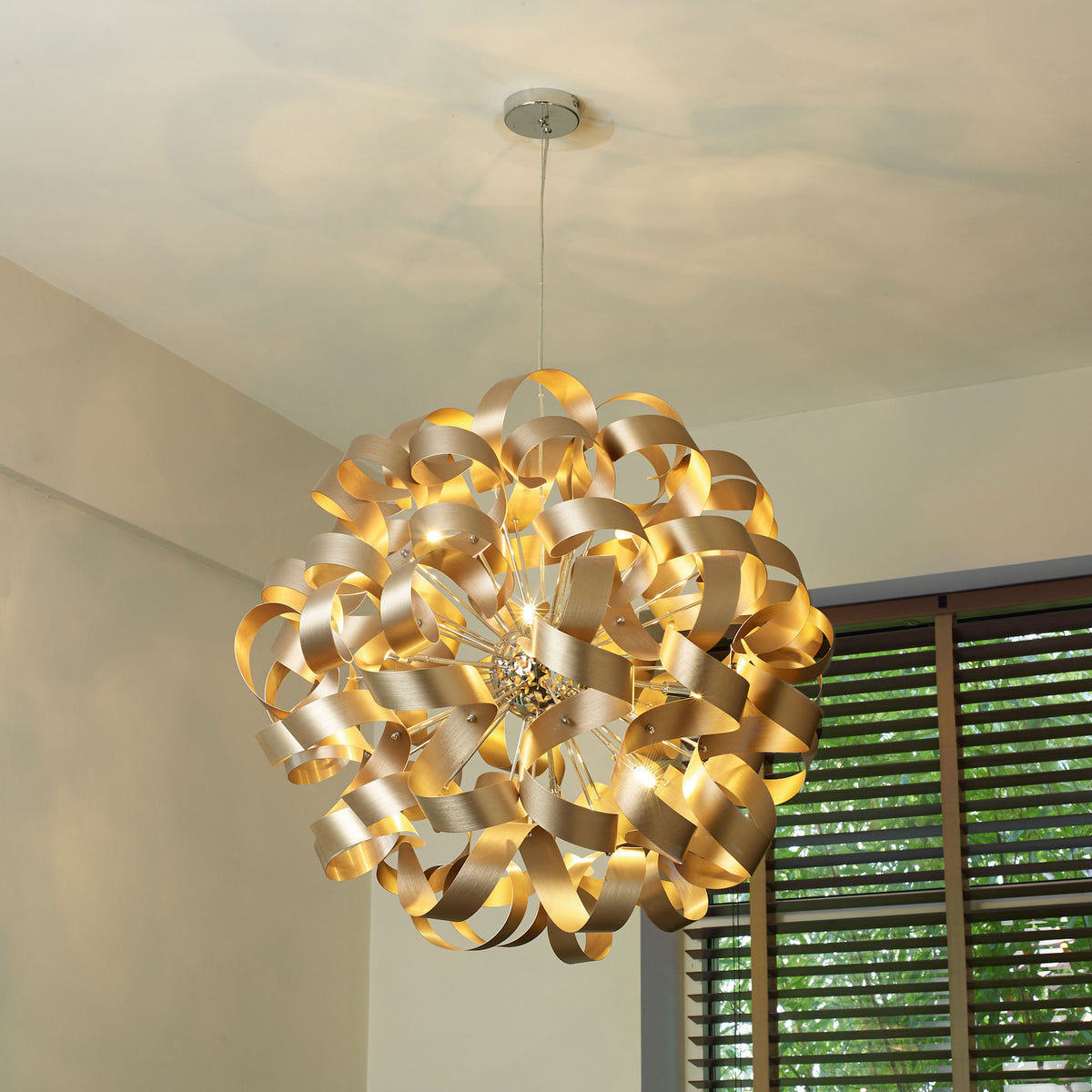 A gold chandelier with swirling metal ribbons hangs from a white ceiling, illuminating a room with warm light.