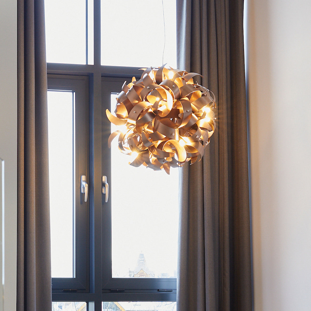 A modern, spherical chandelier with golden, swirling metal strips hangs in front of a window with brown curtains.