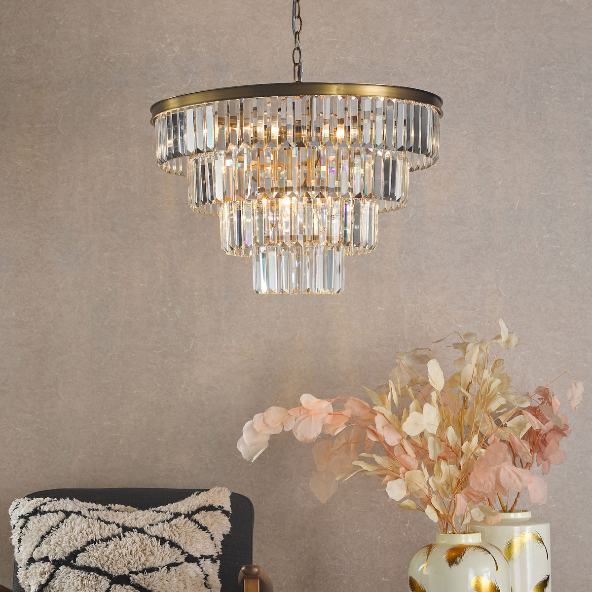 Tiered crystal chandelier with brass accents, illuminating a room with a textured wall, a chair, and decorative vases