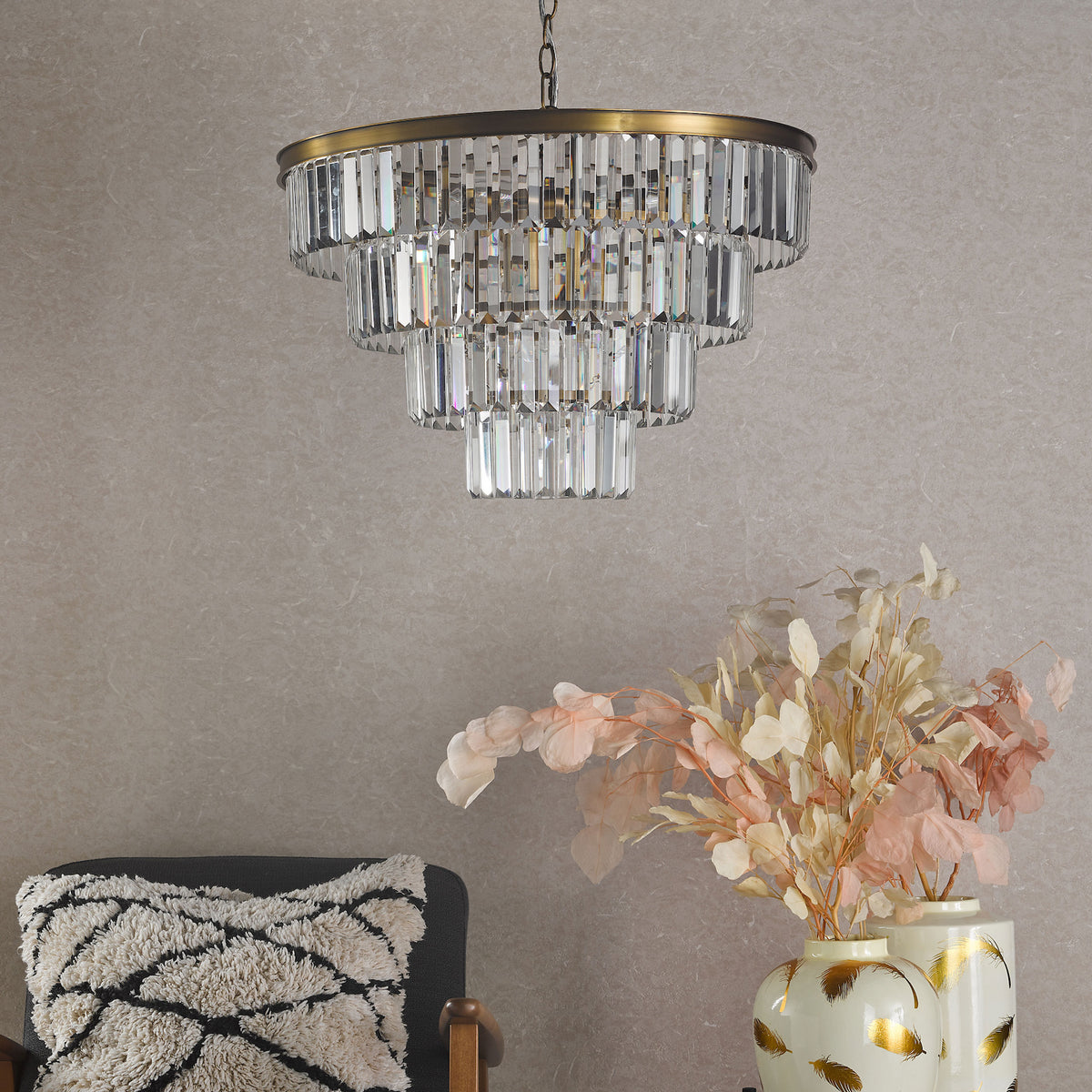 Tiered crystal chandelier with brass accents hanging above a dark chair with a patterned pillow and two vases
