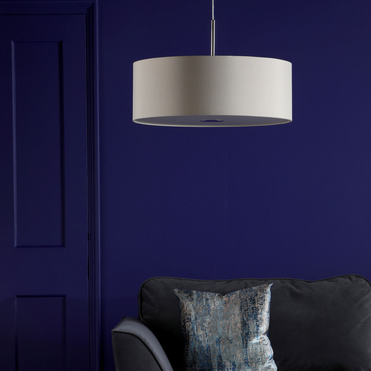 A large, light-colored drum pendant light hangs above a dark gray sofa with a blue and gray patterned pillow.