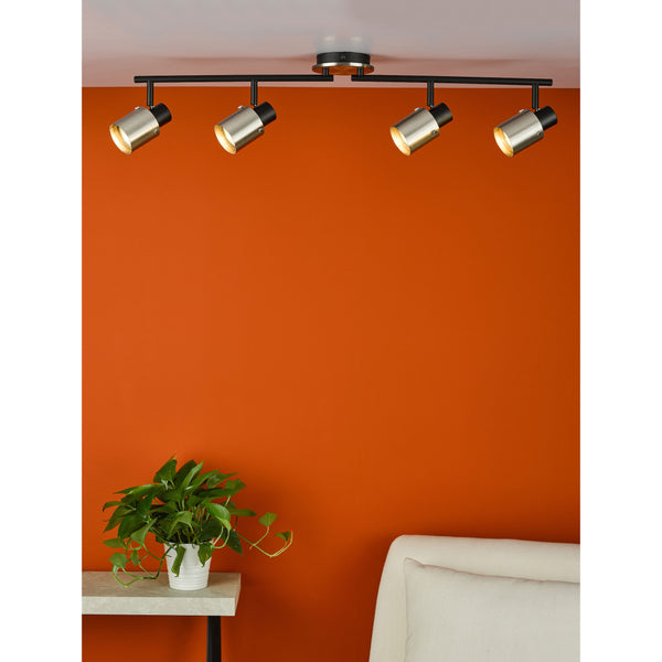Four track lights with brushed metal shades illuminate an orange wall above a plant and a cream sofa.