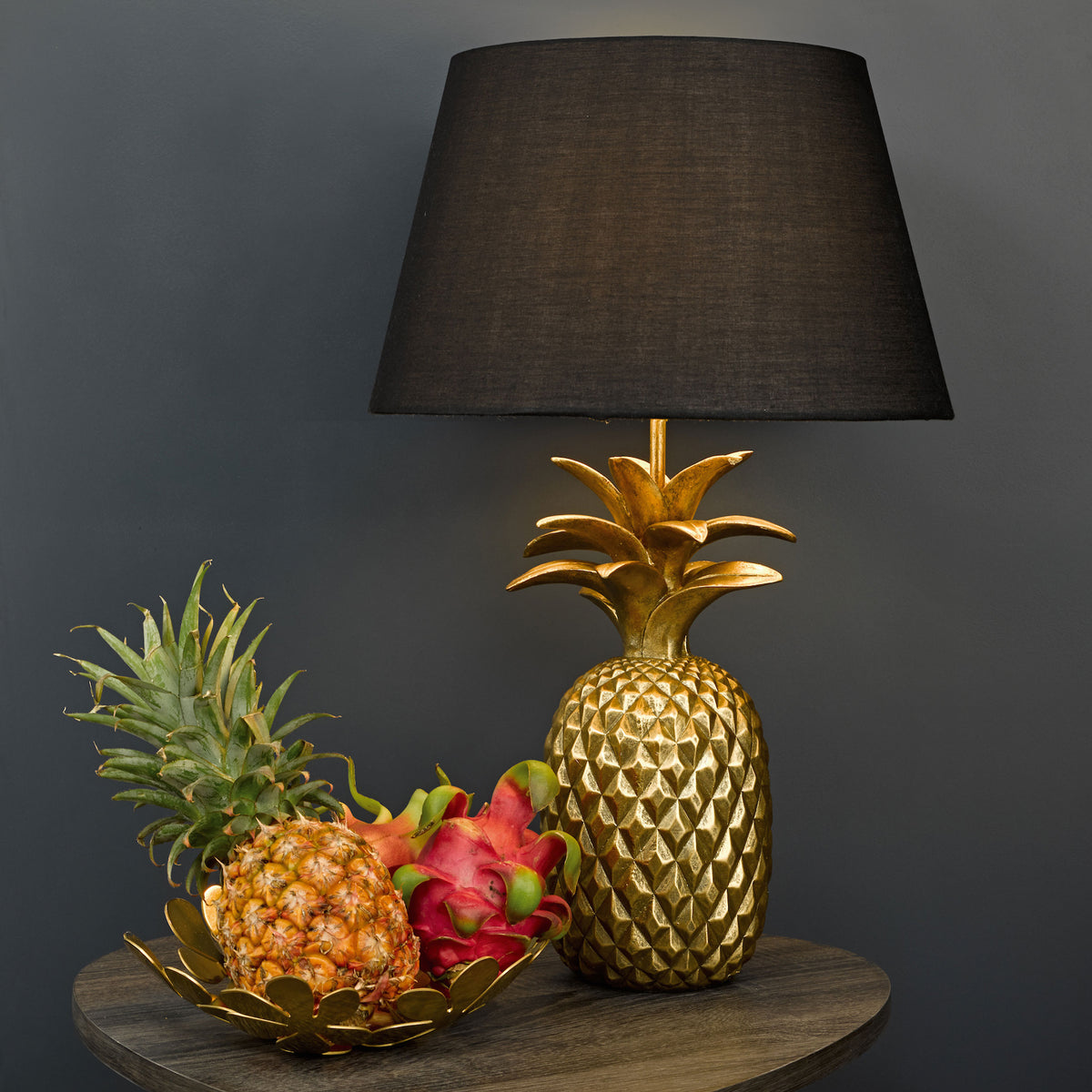 Golden pineapple lamp with a black shade, next to a bowl of pineapples and dragon fruit.