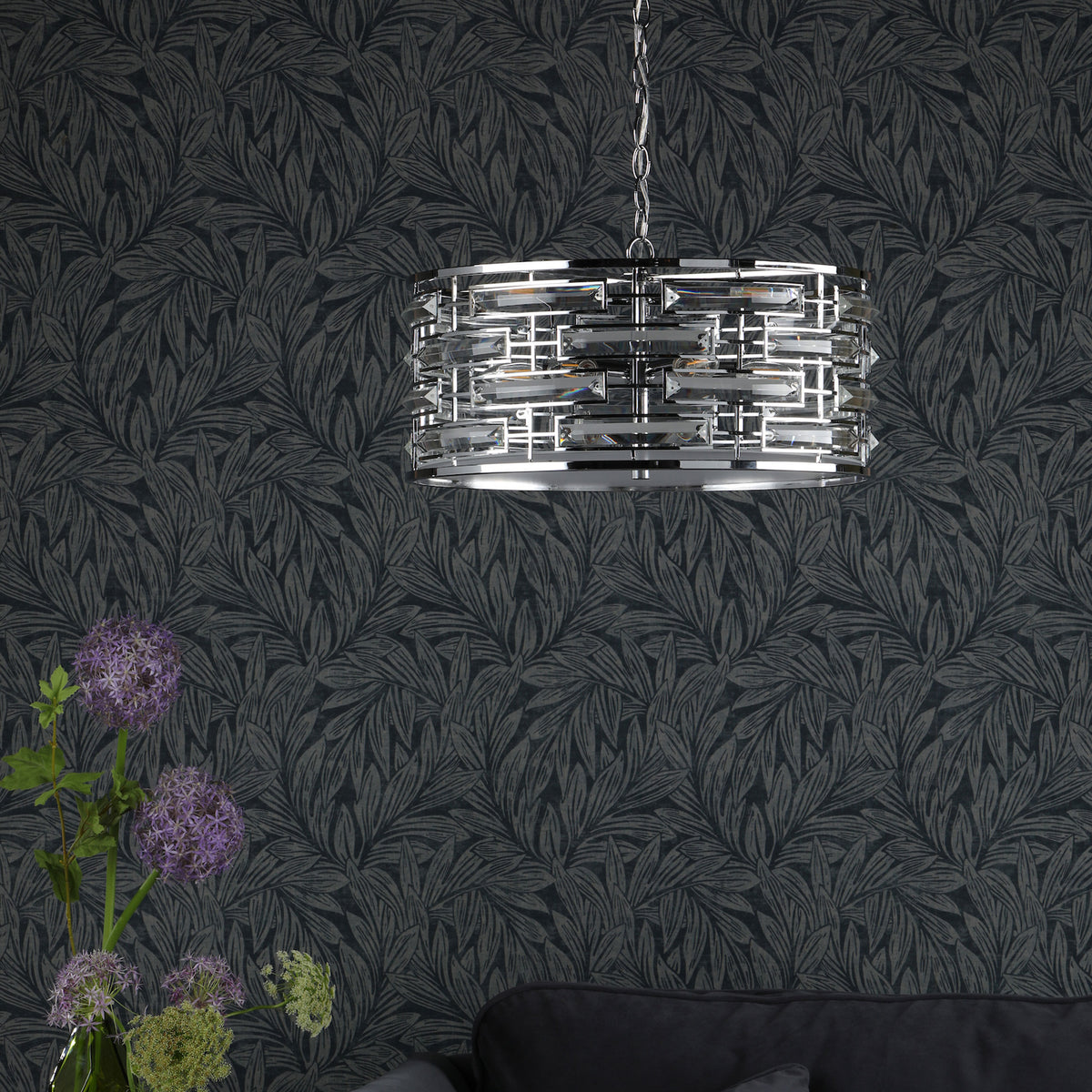 A modern crystal chandelier hangs against dark wallpaper with a subtle leaf pattern. Purple flowers are in a vase.