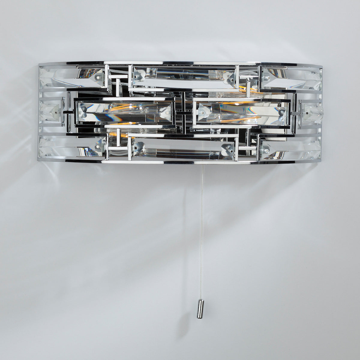 Chrome and crystal wall light with a pull cord switch, casting light on a white wall.
