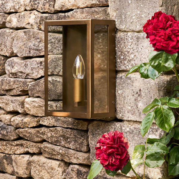 Sandford Outdoor Single Box Lantern in antique brass mounted on a stone wall next to red roses