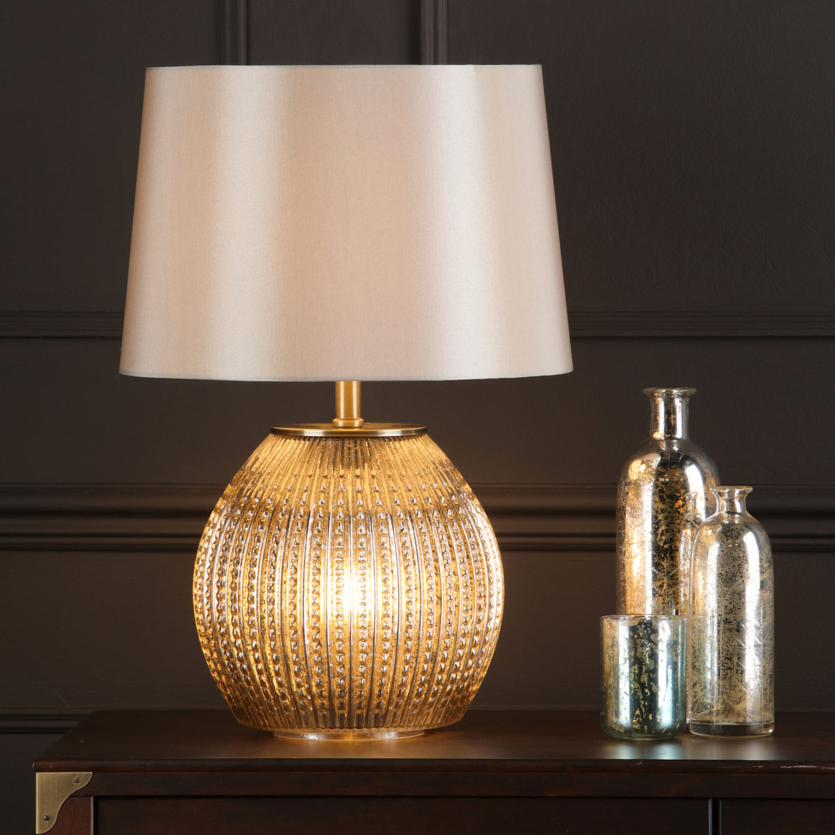 A gold-ribbed glass lamp with a cream shade and two mercury glass bottles on a dark wood table.