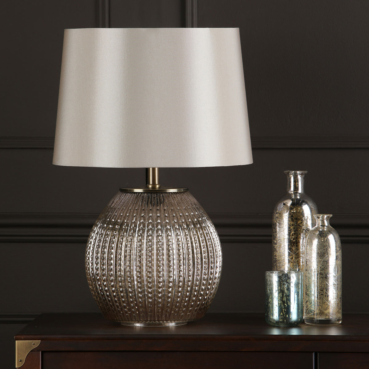 Table lamp with a textured glass base and a light-colored shade, next to decorative mercury glass bottles.