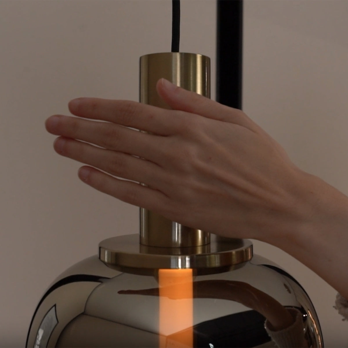 Hand touching a modern pendant light with a brass top and a reflective glass shade, showing a warm glow.