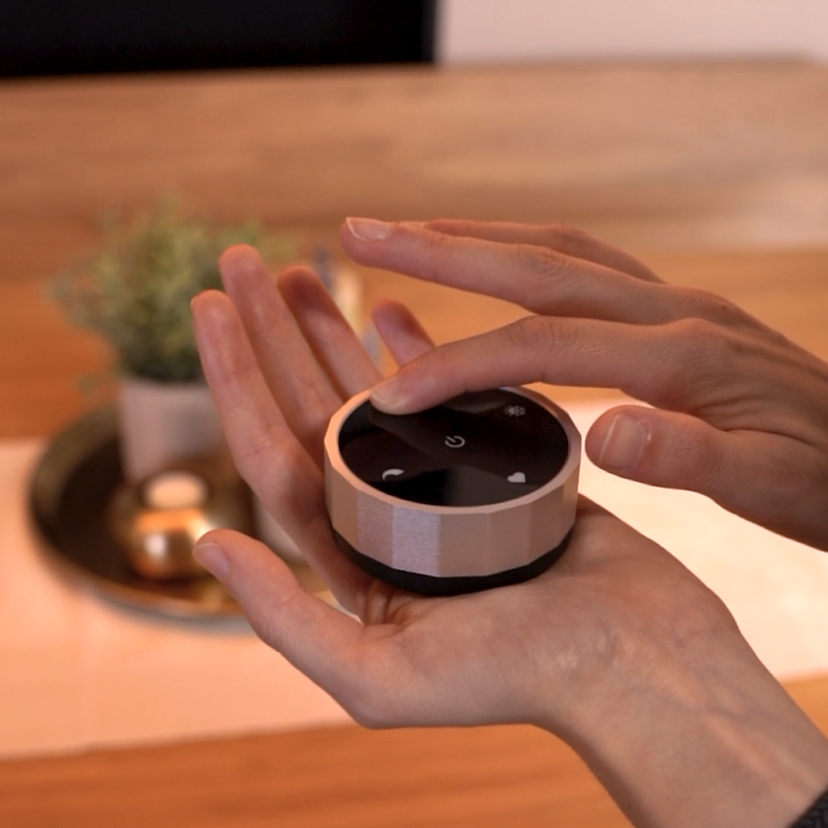Hands holding a small, round, rose gold and black device with a power button and other icons