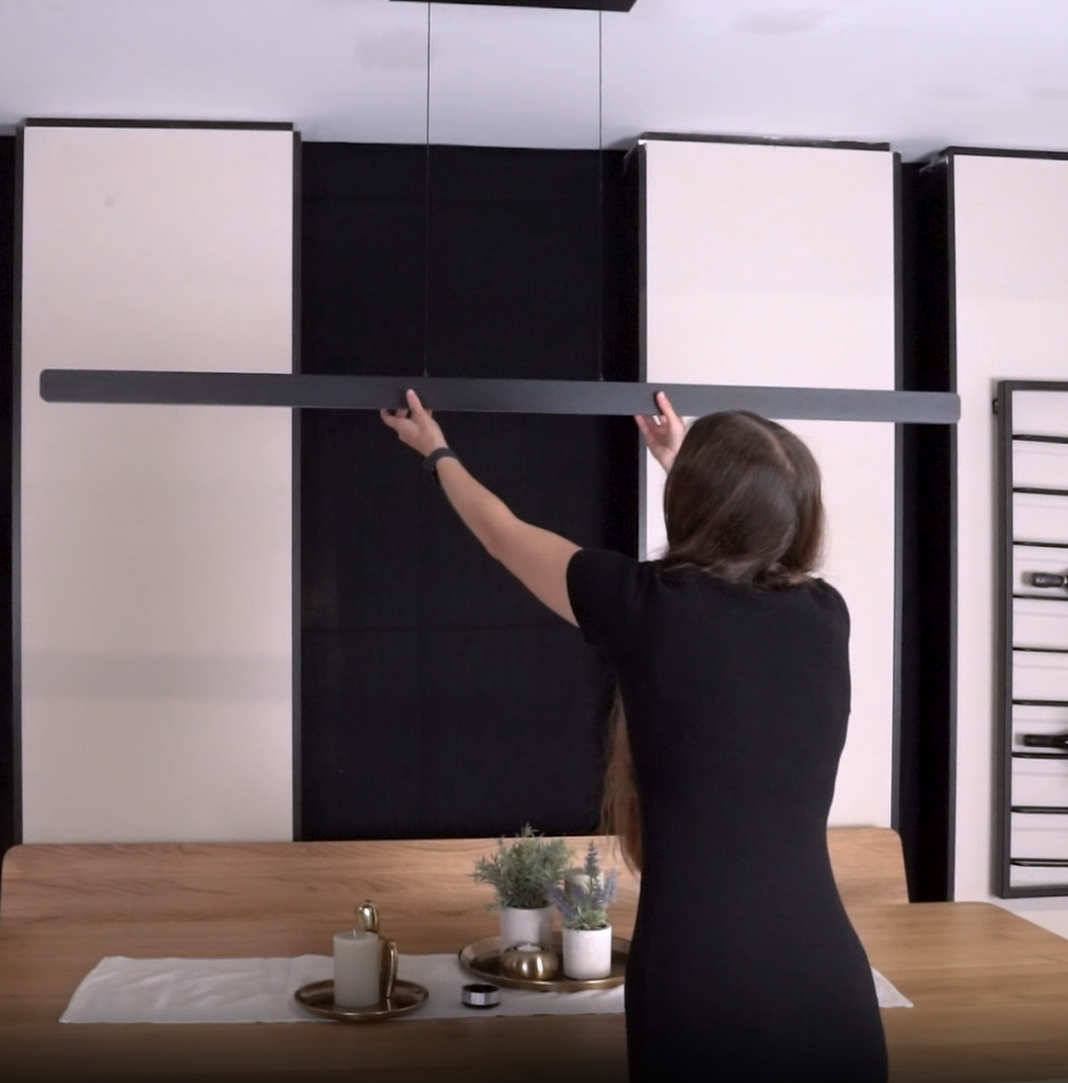 Woman installing a black linear pendant light above a wooden dining table