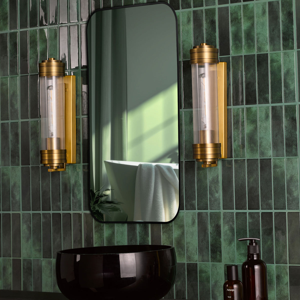 Bathroom with dark green vertical tiles, a black framed mirror, two gold wall sconces, and a black sink