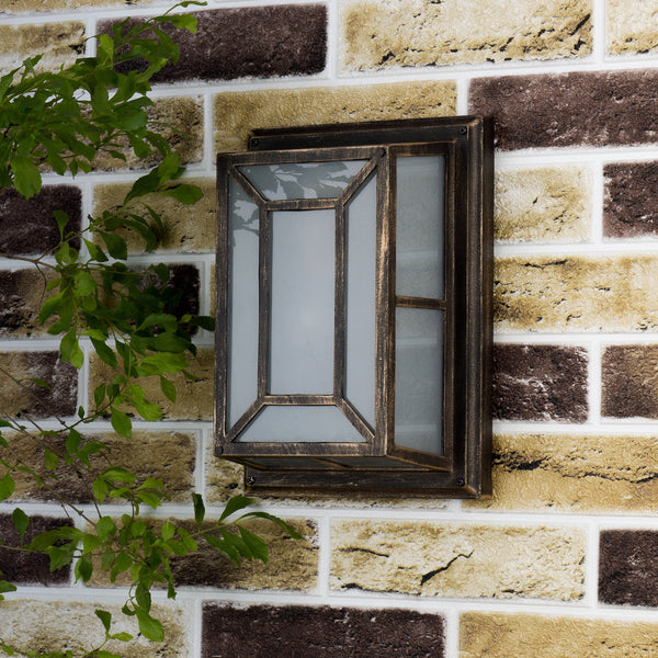 A bronze outdoor wall light with frosted glass panels on a brick wall, with green foliage on the left.