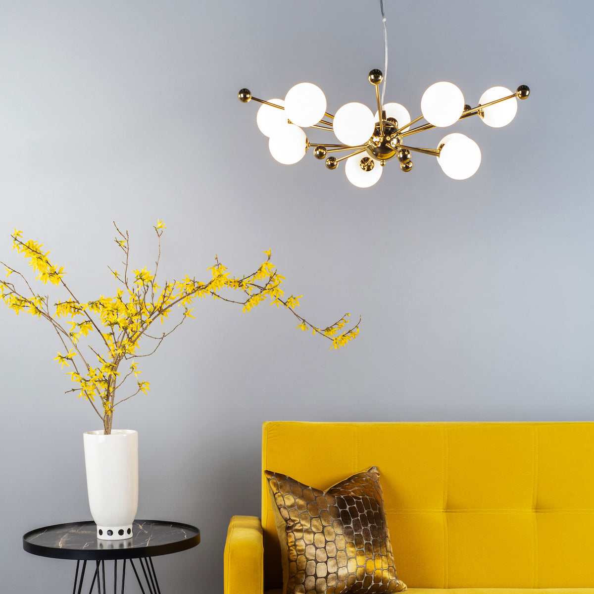 A modern living room with a yellow sofa, a black side table with a white vase of yellow flowers, and a gold chandelier.