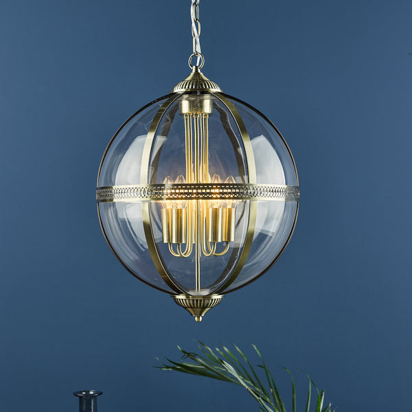 A brass and glass globe chandelier hangs against a deep blue wall, with a green plant in the foreground.